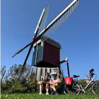Pieterpad Trail cyclists rest beside a traditional windmill, Netherlands.